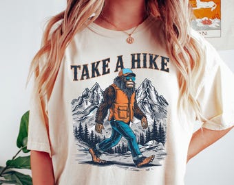 May include: A cream-colored short-sleeve t-shirt featuring a graphic print of a hairy creature wearing a blue cap, sunglasses, and an orange vest. The text "TAKE A HIKE" is printed above the creature, which is walking in front of mountains.