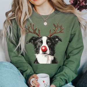 May include: Green sweatshirt featuring a Border Collie with reindeer antlers and a red nose. The dog graphic is centered on the front of the long-sleeved top. The person is wearing blue jeans and holding a white mug with floral designs.