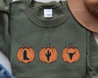 May include: A military green sweatshirt featuring three orange pumpkins with western-themed designs. The pumpkins have a boot, cactus, and bull skull graphic. The text "MILITARY GREEN" is at the bottom. A brown hat and jeans are in the background.