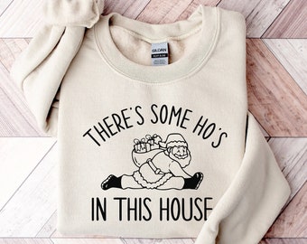 May include: Sand-colored sweatshirt with the text "THERE'S SOME HO'S IN THIS HOUSE" and a graphic of Santa Claus. The Santa illustration is black and white, with Santa holding a sack of gifts. The sweatshirt has a crew neck and long sleeves.