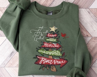 May include: Military green sweatshirt with a Christmas tree design. The tree is made of arrows with text that reads "Wonderful Counselor, Mighty God, Everlasting Father, Prince of Peace." A red cardinal is perched on the tree.