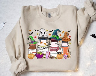 May include: A sand-colored crewneck sweatshirt featuring a graphic print of four medical vials wearing witch hats, surrounded by Halloween elements including pumpkins, a purple cauldron, a small coffin, and bats on a spiderweb background.