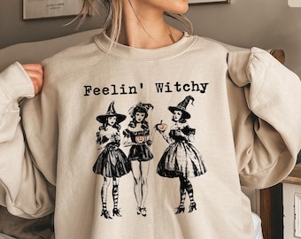 Può includere: Una felpa girocollo color sabbia con una stampa grafica nera di tre figure con cappelli da strega e abiti. Il testo "Feelin' Witchy" è stampato in nero sopra le figure. La felpa ha maniche lunghe e un colletto a coste.