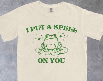 May include: Cream-colored t-shirt with a green graphic of a frog on a lily pad, with the text "I PUT A SPELL ON YOU". The design includes stars and is centered on the shirt. The brand name "TeeShirtApparel" is at the bottom.