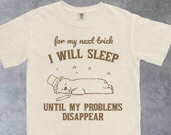 May include: A cream-colored short-sleeve t-shirt featuring brown text that reads for my next trick I WILL SLEEP UNTIL MY PROBLEMS DISAPPEAR. Below the text is a graphic of a bear wearing a top hat, lying down with small stars above it.