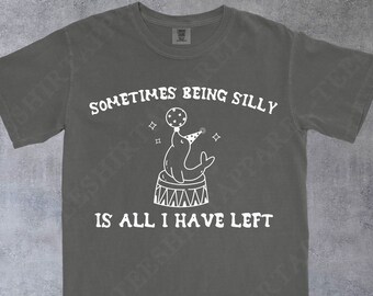 May include: Dark gray t-shirt with the text "SOMETIMES BEING SILLY IS ALL I HAVE LEFT" and a white illustration of a seal balancing a ball on its nose while wearing a party hat. The shirt has a crew neck and short sleeves.
