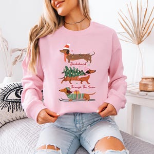 May include: A light pink sweatshirt featuring three dachshunds in festive holiday attire. The top dachshund wears a Santa hat, the middle one carries a Christmas tree, and the bottom one skis. Text reads "Dachshund Through The Snow."