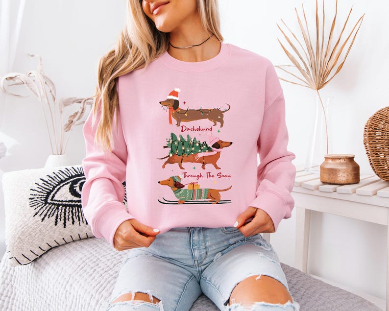 May include: A light pink sweatshirt featuring three dachshunds in festive holiday attire. The top dachshund wears a Santa hat, the middle one carries a Christmas tree, and the bottom one skis. Text reads "Dachshund Through The Snow."