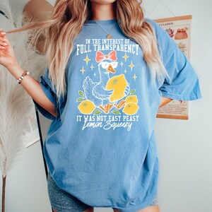 Peut inclure: T-shirt bleu clair avec un dessin d'oie portant des lunettes de soleil et tenant un citron. Le texte sur le t-shirt se lit comme suit : "IN THE INTEREST OF FULL TRANSPARENCY IT WAS NOT EASY PEASY Lemon Squeezy."
