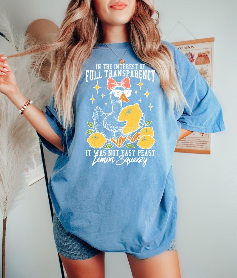 Peut inclure: T-shirt bleu clair avec un dessin d'oie portant des lunettes de soleil et tenant un citron. Le texte sur le t-shirt se lit comme suit : "IN THE INTEREST OF FULL TRANSPARENCY IT WAS NOT EASY PEASY Lemon Squeezy."