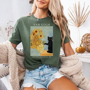 May include: Green t-shirt featuring a print of Van Gogh's "Sunflowers" painting with a black cat. The text "VAN GOGH" is above the image, and "SUNFLOWERS (THIRD VERSION) - 1888" is below. The shirt is worn with denim shorts.