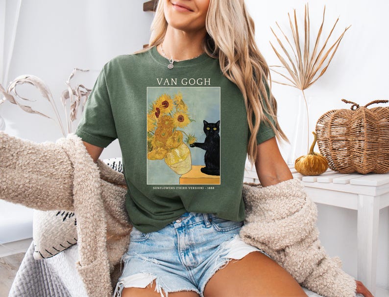 May include: Green t-shirt featuring a print of Van Gogh's "Sunflowers" painting with a black cat. The text "VAN GOGH" is above the image, and "SUNFLOWERS (THIRD VERSION) - 1888" is below. The shirt is worn with denim shorts.
