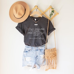 May include: A dark gray t-shirt with the quote "A Dragon without a Rider is a tragedy. A Rider without a Dragon is dead." and a dragonfly graphic. The shirt is paired with denim shorts, a tan hat, and a macrame bag.