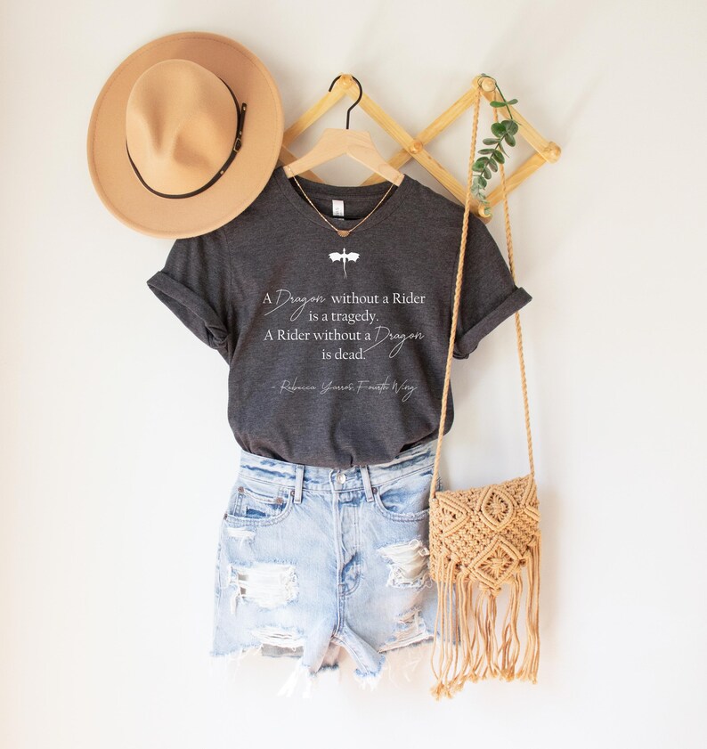 May include: A dark gray t-shirt with the quote "A Dragon without a Rider is a tragedy. A Rider without a Dragon is dead." and a dragonfly graphic. The shirt is paired with denim shorts, a tan hat, and a macrame bag.