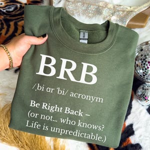 May include: An olive green sweatshirt with the acronym "BRB" in large white letters. Below the acronym is the definition: "/bi ar 'bi/ acronym Be Right Back – (or not... who knows? Life is unpredictable.)"