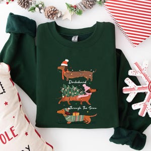 May include: A dark green sweatshirt featuring three dachshunds, each adorned with festive holiday accessories. The text "Dachshund Through The Snow" is printed on the shirt. A Christmas-themed pillow and snowflake are in the background.