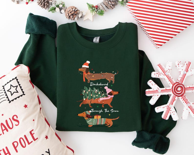 May include: A dark green sweatshirt featuring three dachshunds, each adorned with festive holiday accessories. The text "Dachshund Through The Snow" is printed on the shirt. A Christmas-themed pillow and snowflake are in the background.