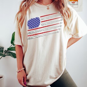 K&ouml;nnte beinhalten: Cremefarbenes T-Shirt mit einem stilisierten Design der amerikanischen Flagge. Die Flagge enth&auml;lt ein blaues Sternenfeld und rot-wei&szlig;e Streifen, mit D&uuml;senjets, die Kondensstreifen hinterlassen. Der Text "TeeShirtApparel" befindet sich unten.