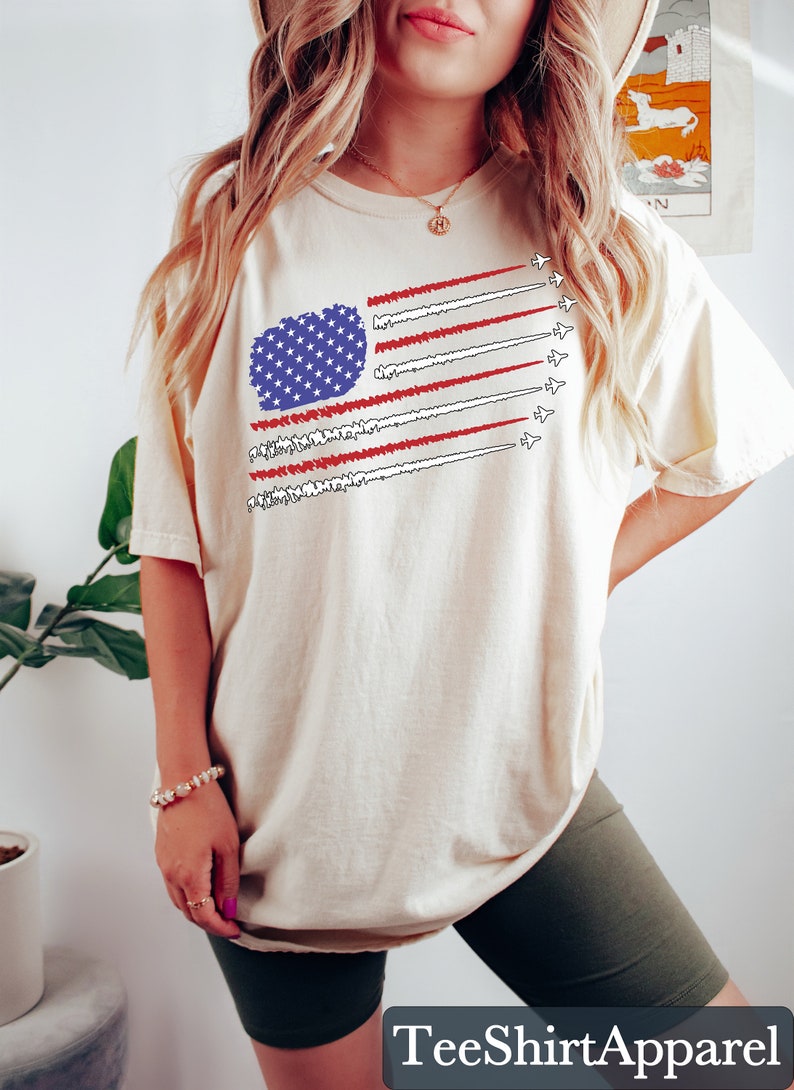 K&ouml;nnte beinhalten: Cremefarbenes T-Shirt mit einem stilisierten Design der amerikanischen Flagge. Die Flagge enth&auml;lt ein blaues Sternenfeld und rot-wei&szlig;e Streifen, mit D&uuml;senjets, die Kondensstreifen hinterlassen. Der Text "TeeShirtApparel" befindet sich unten.