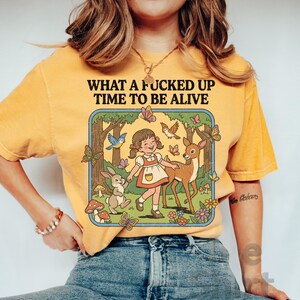 May include: Mustard-colored t-shirt with the text "WHAT A FUCKED UP TIME TO BE ALIVE." The graphic features a cartoon girl, deer, rabbit, butterflies, and birds in a forest scene. The shirt is made of a soft material.