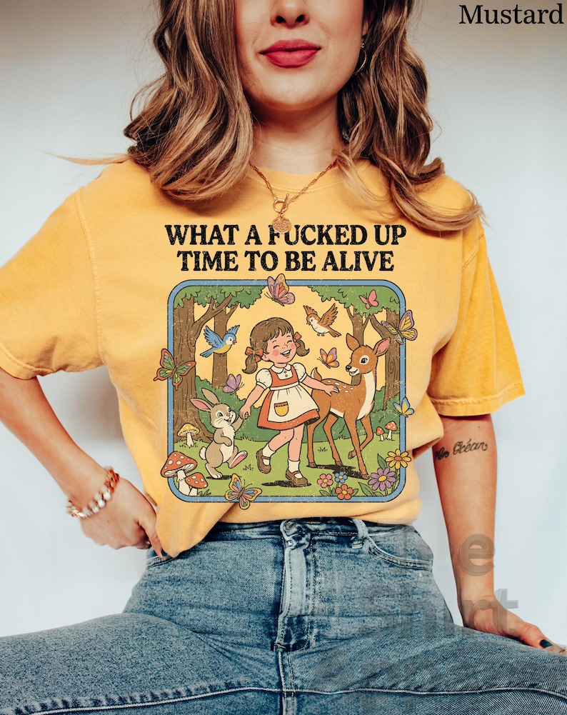 May include: Mustard-colored t-shirt with the text "WHAT A FUCKED UP TIME TO BE ALIVE." The graphic features a cartoon girl, deer, rabbit, butterflies, and birds in a forest scene. The shirt is made of a soft material.