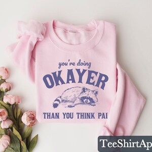 Puede incluir: Sudadera rosa claro con la frase "you're doing OKAYER THAN YOU THINK PAI" en azul oscuro. Debajo, una ilustraci&oacute;n detallada de un mapache. La sudadera est&aacute; doblada y se muestra sobre una superficie blanca.