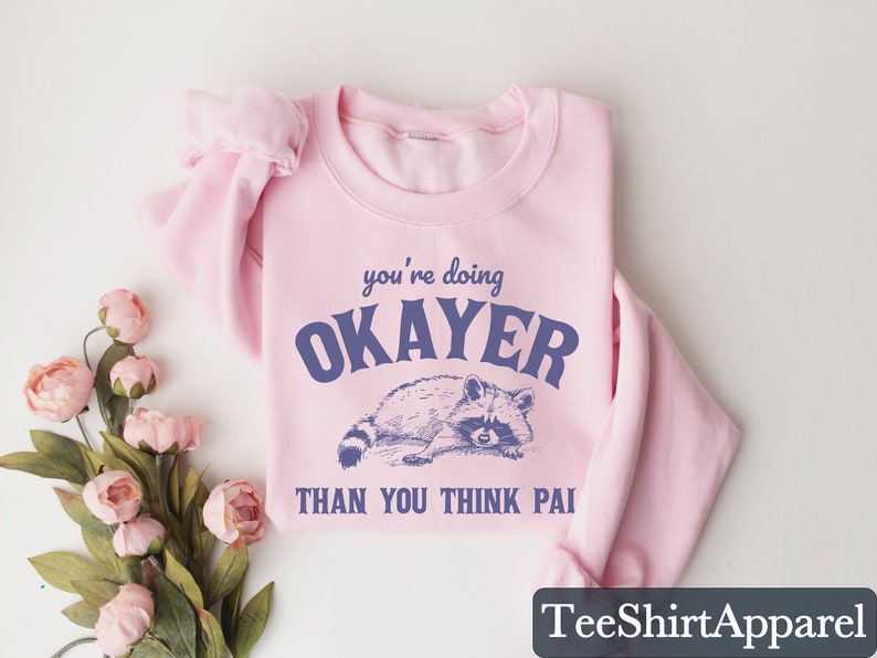 Puede incluir: Sudadera rosa claro con la frase "you're doing OKAYER THAN YOU THINK PAI" en azul oscuro. Debajo, una ilustraci&oacute;n detallada de un mapache. La sudadera est&aacute; doblada y se muestra sobre una superficie blanca.