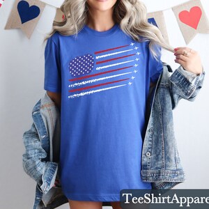 K&ouml;nnte beinhalten: Ein blaues T-Shirt mit einer Grafik der amerikanischen Flagge, mit wei&szlig;en und roten Streifen und einem Sternenkreis. Das Shirt hat einen Rundhalsausschnitt und kurze &Auml;rmel. Der Text "TeeShirtApparel" befindet sich unten.