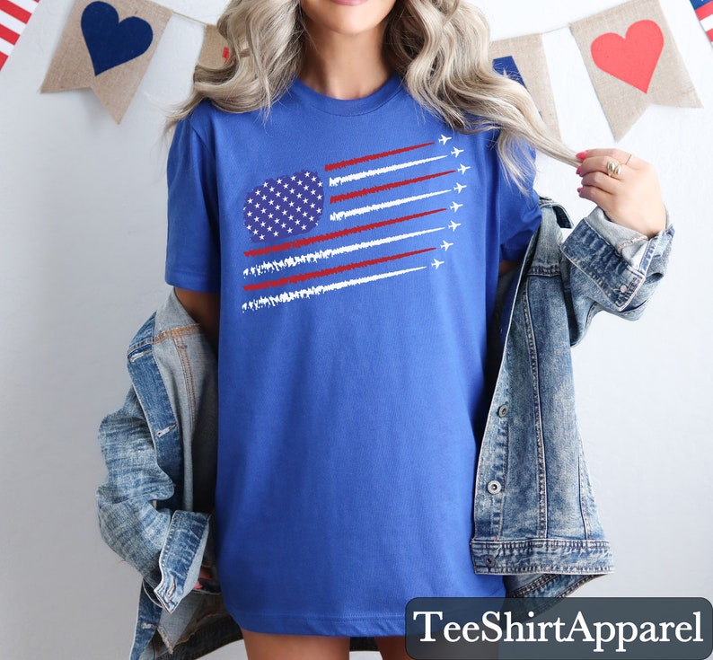 K&ouml;nnte beinhalten: Ein blaues T-Shirt mit einer Grafik der amerikanischen Flagge, mit wei&szlig;en und roten Streifen und einem Sternenkreis. Das Shirt hat einen Rundhalsausschnitt und kurze &Auml;rmel. Der Text "TeeShirtApparel" befindet sich unten.