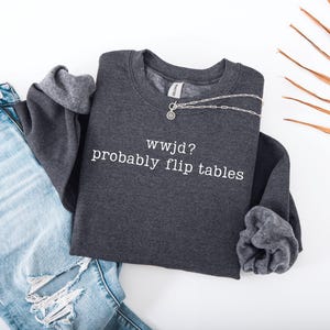 Peut inclure: Un sweat-shirt gris fonc&eacute; avec le texte blanc "wwjd? probably flip tables". Le sweat-shirt est associ&eacute; &agrave; un jean en denim bleu clair avec des d&eacute;tails d&eacute;chir&eacute;s. Un collier argent&eacute; avec un petit pendentif est sur le sweat-shirt.