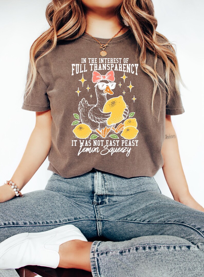 Peut inclure: T-shirt marron avec un dessin d'oie portant des lunettes de soleil et un n&oelig;ud, tenant un citron. Le texte dit "IN THE INTEREST OF FULL TRANSPARENCY" et "IT WAS NOT EASY PEASY Lemon Squeezy".