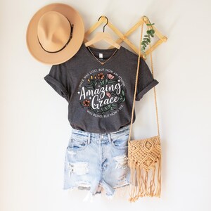 May include: A dark gray t-shirt with the text "Amazing Grace" surrounded by floral designs. The shirt is paired with denim shorts, a tan hat, and a woven crossbody bag. The text on the shirt reads "I once was lost, but now am found. Was blind, but now I see."