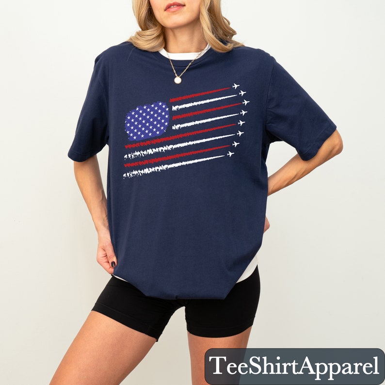 K&ouml;nnte beinhalten: Marineblaues T-Shirt mit einem stilisierten Design der amerikanischen Flagge, das ein blaues Oval mit wei&szlig;en Sternen und rot-wei&szlig;en Streifen mit wei&szlig;en Flugzeugspuren zeigt. Der Text "TeeShirtApparel" befindet sich unten.