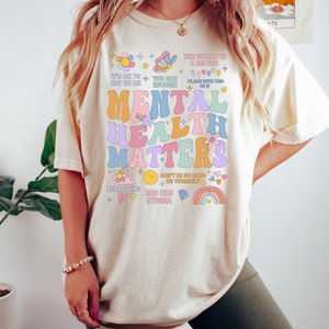 May include: Cream-colored t-shirt with the text "MENTAL HEALTH MATTERS" in colorful, retro-style lettering. The shirt includes positive affirmations and graphics like flowers, rainbows, and stars. The brand name "TeeShirtApparel" is at the bottom.