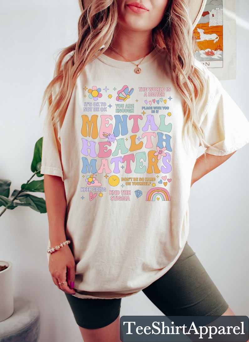 May include: Cream-colored t-shirt with the text "MENTAL HEALTH MATTERS" in colorful, retro-style lettering. The shirt includes positive affirmations and graphics like flowers, rainbows, and stars. The brand name "TeeShirtApparel" is at the bottom.