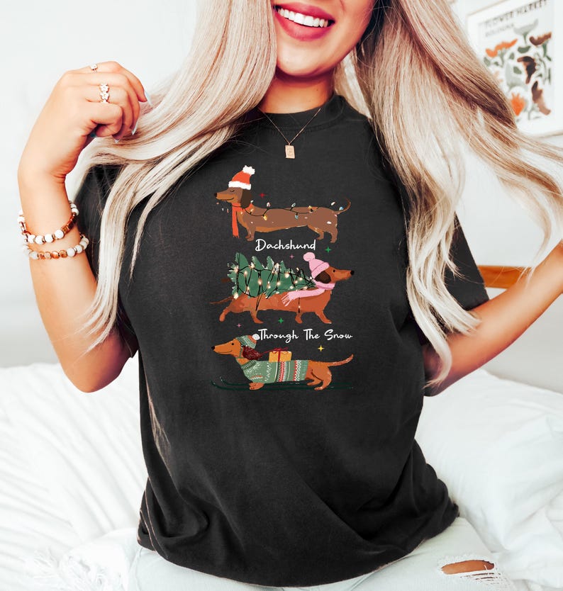 May include: Black t-shirt featuring three dachshunds in festive holiday attire. The dogs are depicted with a Santa hat, carrying a Christmas tree, and wearing a sweater. The text "Dachshund Through The Snow" is also on the shirt.