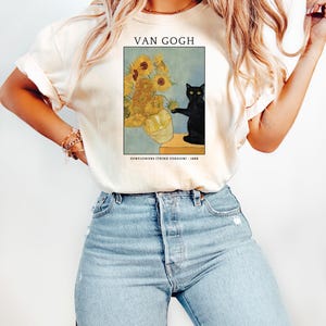 May include: Cream-colored t-shirt featuring a framed print of Van Gogh's "Sunflowers (Third Version)" with a black cat. The print includes the text "VAN GOGH" and "SUNFLOWERS (THIRD VERSION) - 1888". The shirt is paired with blue jeans.