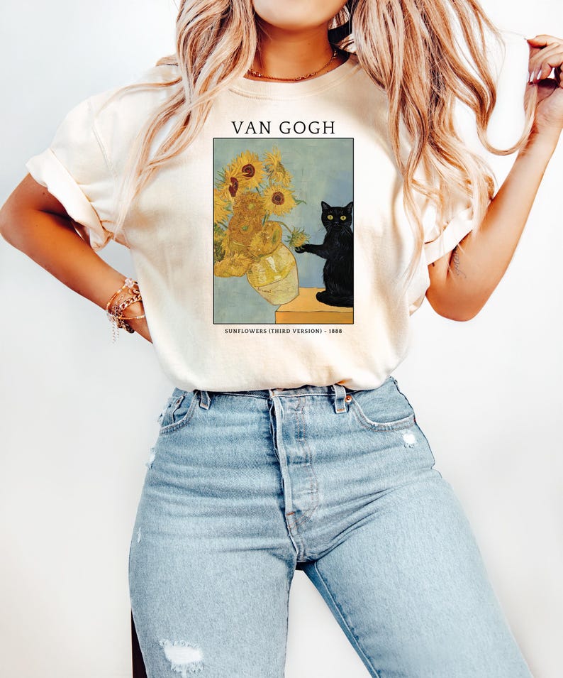 May include: Cream-colored t-shirt featuring a framed print of Van Gogh's "Sunflowers (Third Version)" with a black cat. The print includes the text "VAN GOGH" and "SUNFLOWERS (THIRD VERSION) - 1888". The shirt is paired with blue jeans.