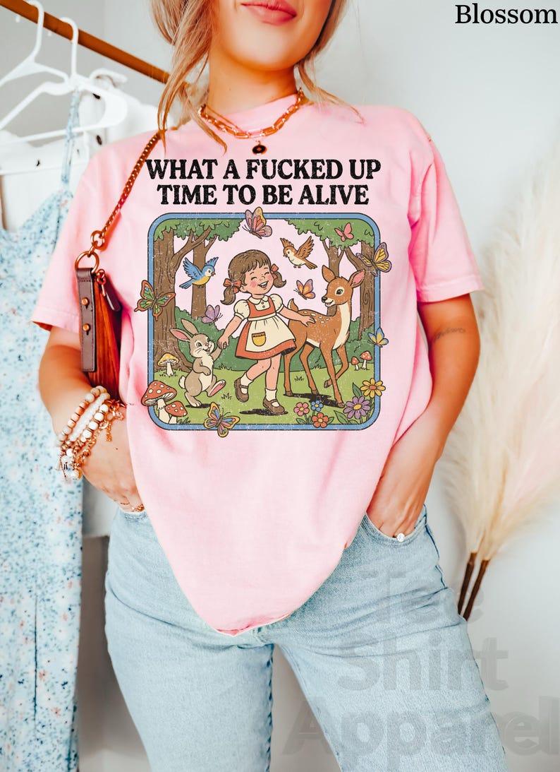 May include: Pink t-shirt with the text "WHAT A FUCKED UP TIME TO BE ALIVE" above a cartoon illustration of a girl, deer, rabbit, and butterflies in a forest scene. The shirt is a light pink color.
