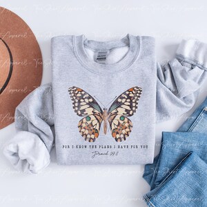 May include: A light gray sweatshirt featuring a detailed butterfly design with brown, cream, and teal accents. The text "FOR I KNOW THE PLANS I HAVE FOR YOU Jeremiah 29:11" is printed below the butterfly. A brown hat and blue jeans are also in the image.