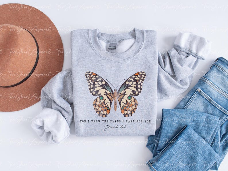 May include: A light gray sweatshirt featuring a detailed butterfly design with brown, cream, and teal accents. The text "FOR I KNOW THE PLANS I HAVE FOR YOU Jeremiah 29:11" is printed below the butterfly. A brown hat and blue jeans are also in the image.