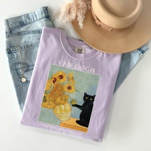 May include: A light purple t-shirt featuring a print of Van Gogh's "Sunflowers" painting with a black cat. The shirt is paired with light wash jeans, a tan hat, and a gold necklace. The text "VAN GOGH" is printed above the artwork.