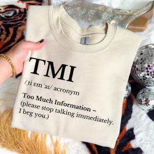 May include: A cream-colored sweatshirt with the acronym "TMI" printed in large, black letters. The text below defines "TMI" as "Too Much Information" and includes a plea to stop talking. The sweatshirt is held by a person.