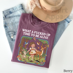 May include: Berry-colored t-shirt with the text "WHAT A FUCKED UP TIME TO BE ALIVE". The graphic features a cartoon girl, deer, rabbit, butterflies, and birds in a forest scene. The shirt is laid on top of blue jeans and a tan hat.