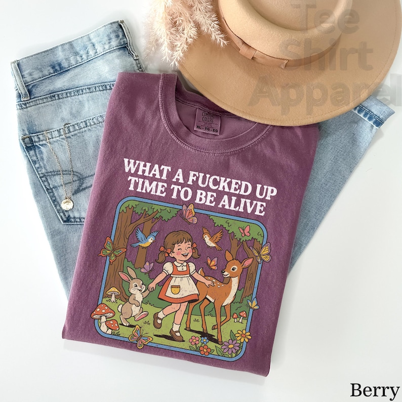 May include: Berry-colored t-shirt with the text "WHAT A FUCKED UP TIME TO BE ALIVE". The graphic features a cartoon girl, deer, rabbit, butterflies, and birds in a forest scene. The shirt is laid on top of blue jeans and a tan hat.