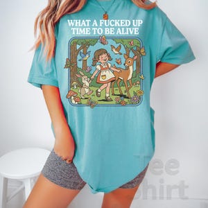 May include: A turquoise t-shirt with the text "WHAT A FUCKED UP TIME TO BE ALIVE" above a cartoon illustration of a girl, deer, rabbit, butterflies, and birds in a forest scene. The shirt is a light, solid color.