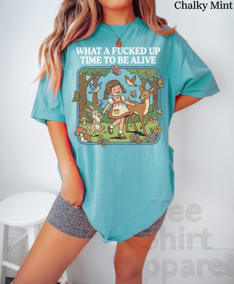 May include: A turquoise t-shirt with the text "WHAT A FUCKED UP TIME TO BE ALIVE" above a cartoon illustration of a girl, deer, rabbit, butterflies, and birds in a forest scene. The shirt is a light, solid color.