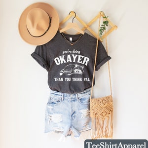 Puede incluir: Camiseta gris oscuro con el texto "you're doing OKAYER than you think pal" y un gr&aacute;fico de mapache. La camiseta se combina con pantalones cortos de mezclilla, un sombrero beige y un bolso cruzado de macram&eacute;. Tambi&eacute;n se ven una percha de madera y decoraci&oacute;n de pared.