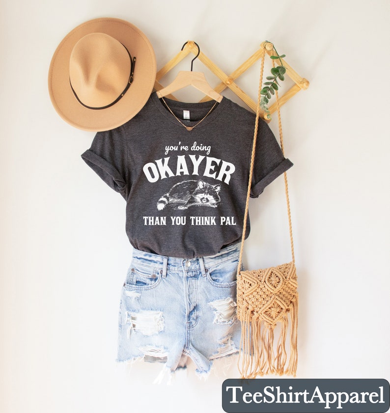 Puede incluir: Camiseta gris oscuro con el texto "you're doing OKAYER than you think pal" y un gr&aacute;fico de mapache. La camiseta se combina con pantalones cortos de mezclilla, un sombrero beige y un bolso cruzado de macram&eacute;. Tambi&eacute;n se ven una percha de madera y decoraci&oacute;n de pared.