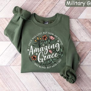 May include: A military green sweatshirt with the text "Amazing Grace" in a floral design. The text "I once was lost, but now am found, was blind, but now I see" is also included. The sweatshirt has long sleeves.